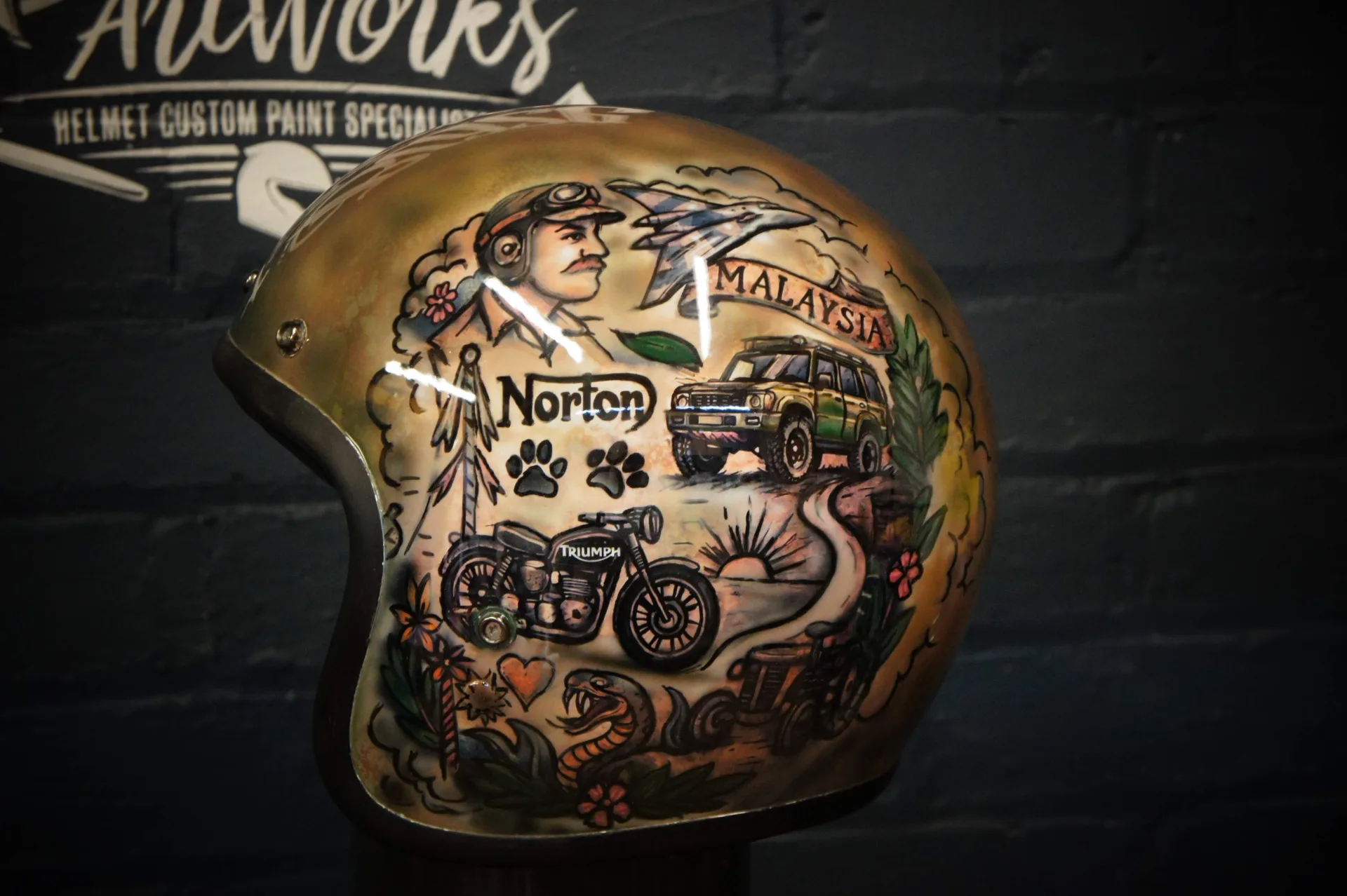 Finished Bell 500 custom helmet, hand-painted in traditional tattoo style on an aged-gold base, featuring an aviator portrait, motorcycles, a fighter jet and floral detail.