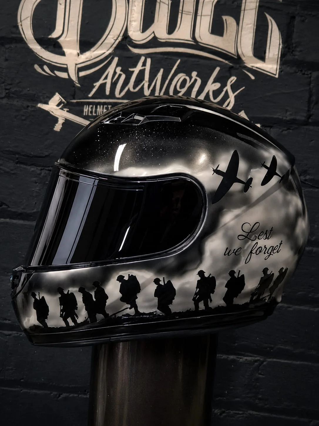 Finished remembrance helmet on a black base, covered in airbrushed stars with marching soldiers, aircraft and 'Lest we forget' lettering.